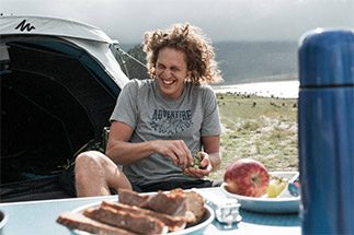 Comer durante um trekking: Rapaz sorrindo em frente à sua barraca enquanto se alimenta com frutas e pães