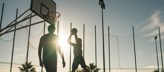 Dois amigos jogando basquete em quadra próxima à praia com por do sol ao fundo
